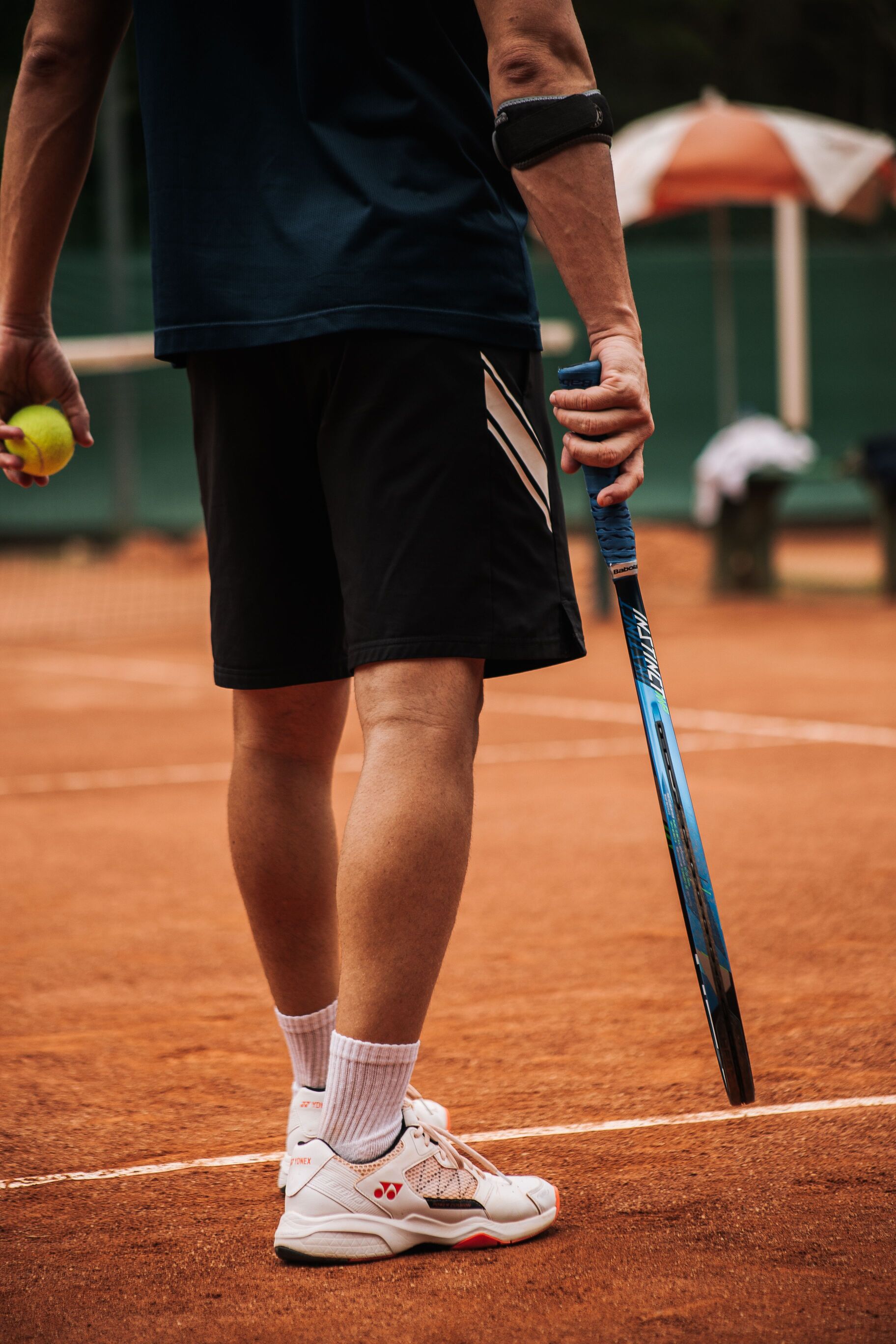 tennis player standing on a clay court holding a racket and a ball, wearing an elbow strap