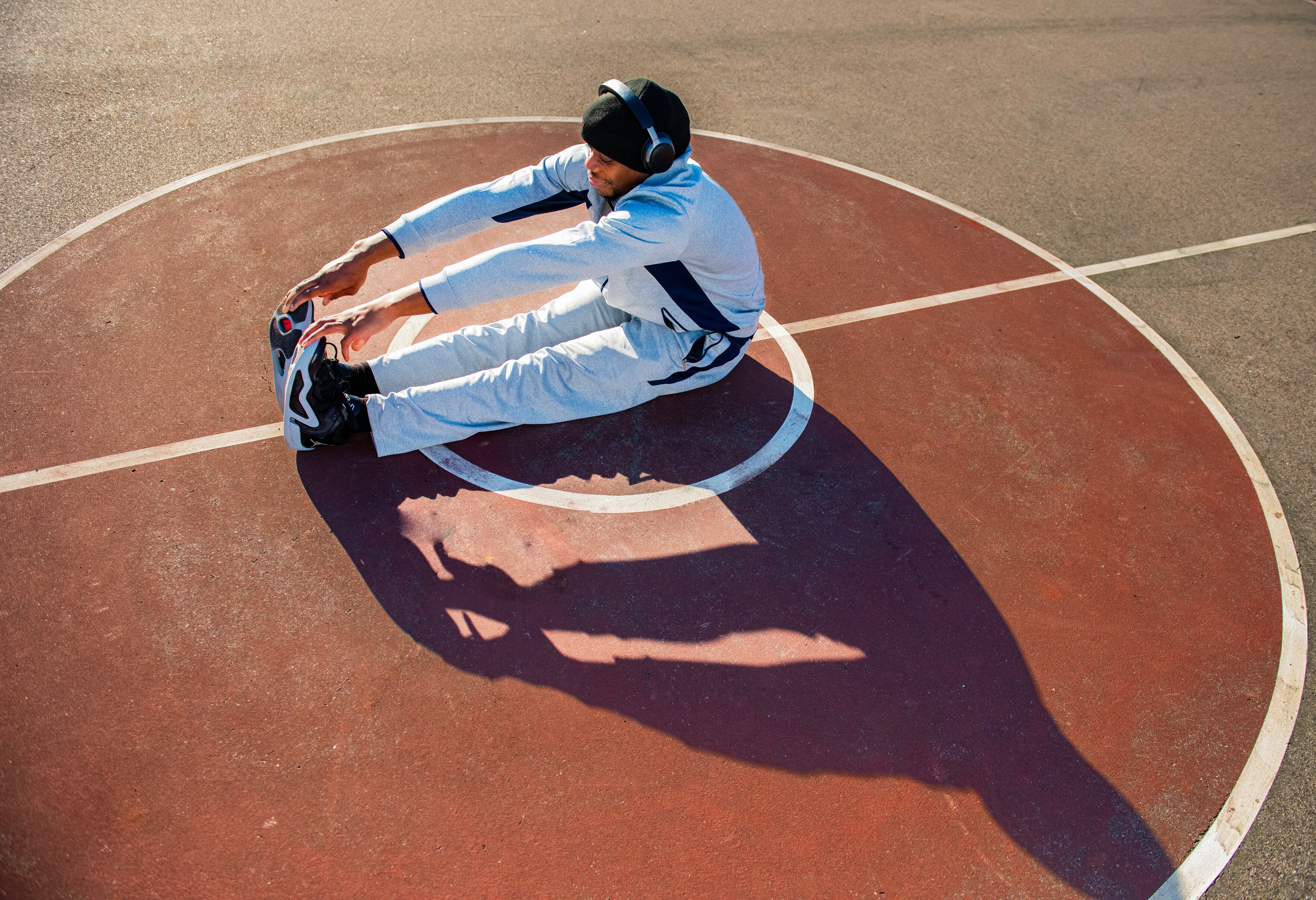 man in a light grey tracksuit and headphones doing a seated forward stretch on a red outdoor court