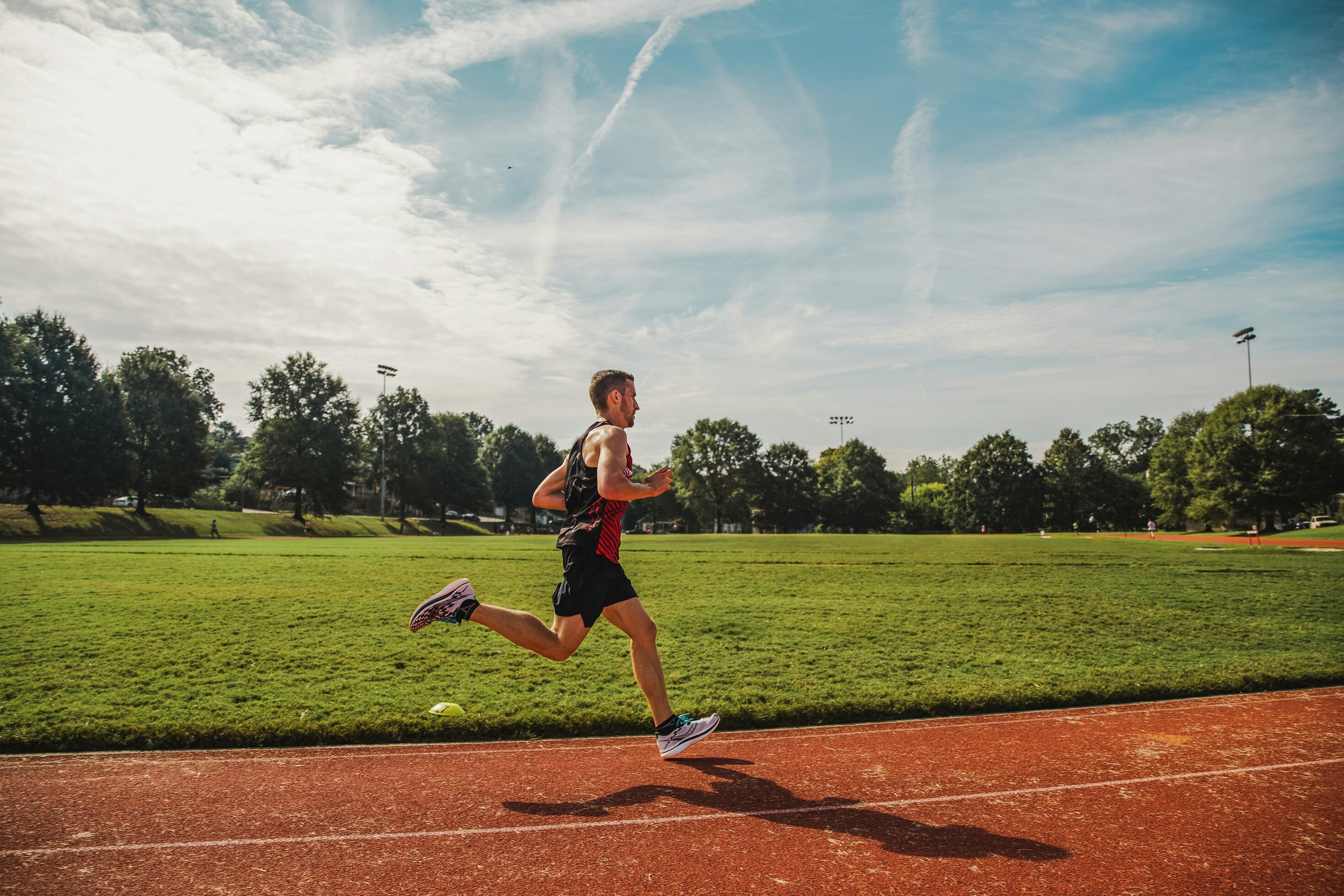 man running on a red track beside a green field under a bright sky with soft clouds